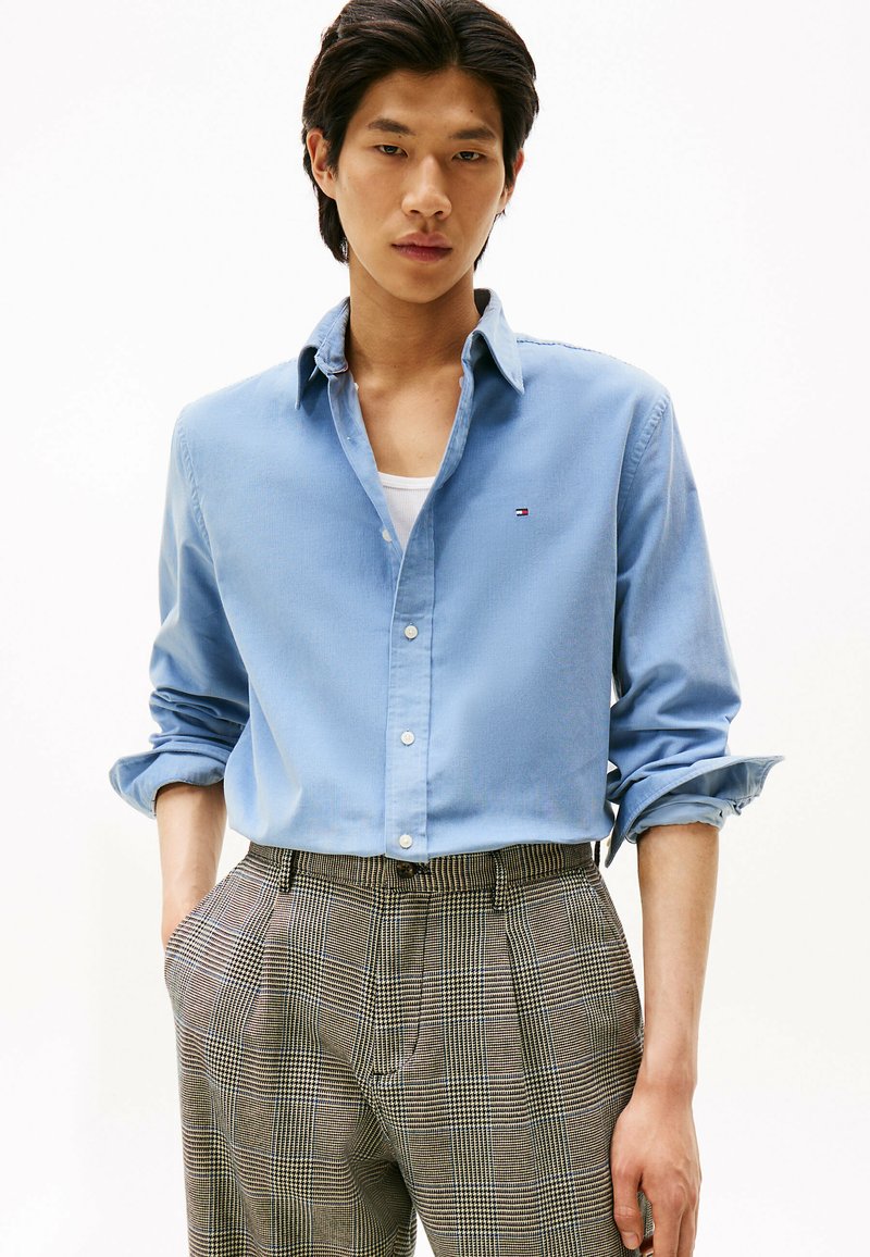 Lichtblauw overhemd met opgerolde mouwen, gedragen over een wit shirt, gecombineerd met bruin geruite high-waisted broek.