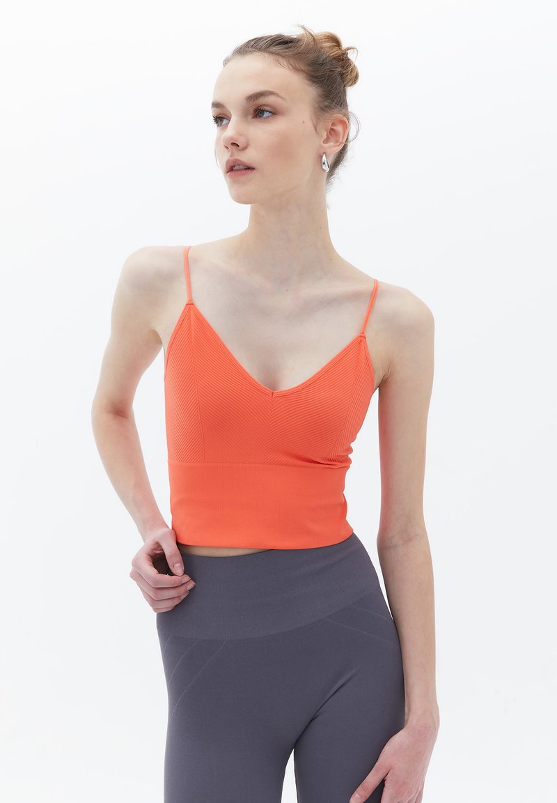 Top crop arancione a coste con scollatura a V, spalline sottili e design aderente, abbinato a leggings grigi a vita alta. Tessuto liscio e silhouette moderna.