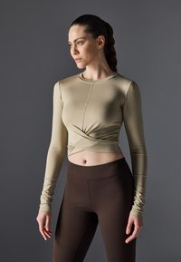 Beige langermet tettsittende topp med vridd front, sammen med mørkebrune høytaljede leggings laget av glatt, elastisk stoff.