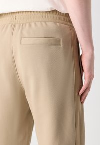 Beige treningsbukser har en teksturert livlinje, en enkelt baklomme og sidesømmer, noe som fremhever et glatt, tettsittende design.