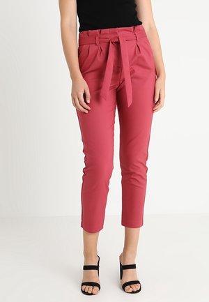 Vrouw draagt hoge, cropped roze broek met een taillekoord, gecombineerd met een zwarte mouwloze top en zwarte sandalen met hak.