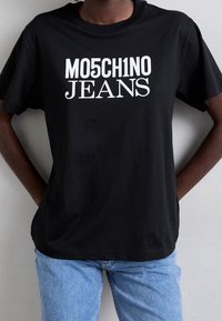 Osoba v černém tričku s nápisem "MO5CH1NO JEANS" vytištěným tučnými bílými písmeny, stojící před světle šedou zdí.