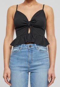 Black ribbed crop top with adjustable spaghetti straps, front tie detail, and a peplum hem, paired with light blue high-waisted jeans.