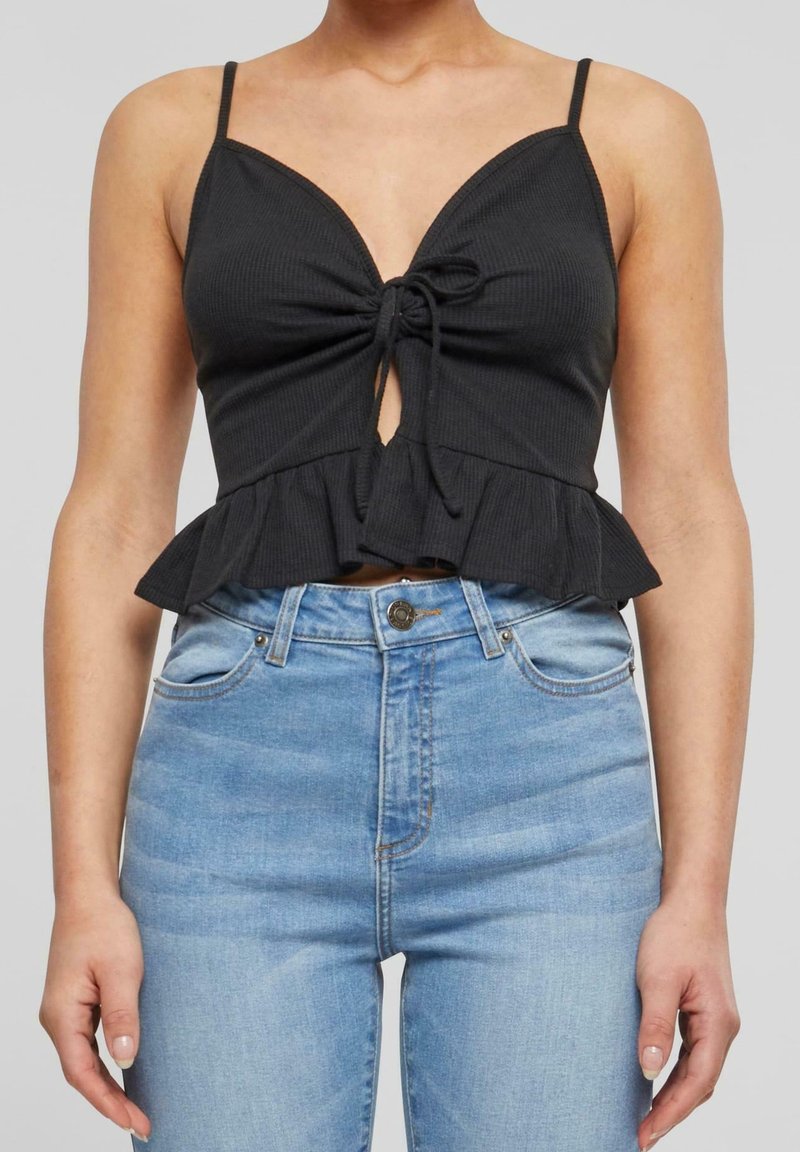 Black ribbed crop top with adjustable spaghetti straps, front tie detail, and a peplum hem, paired with light blue high-waisted jeans.