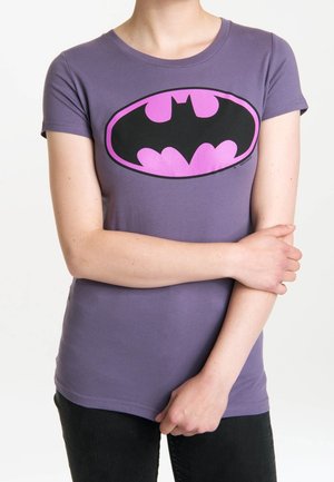 Personne portant un t-shirt ajusté violet avec un logo Batman noir et rose, bras croisés, et pantalon noir visible de la taille vers le bas.
