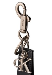 Zilverkleurige metalen sleutelhanger met gegraveerde tekst "Calvin Klein", bevestigd aan een zwarte leren band die een zilveren "cK" logo bedel vasthoudt.