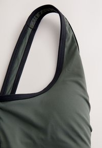 Primer plano de la parte superior de un traje de baño verde oscuro con ribete negro en la tira y el escote sobre un fondo claro.