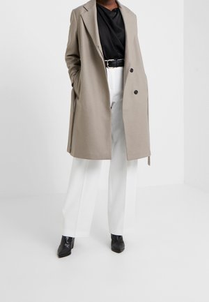 Personne portant un long manteau beige, un haut noir, un pantalon blanc, une ceinture noire et des bottes noires pointues, debout les mains dans les poches.