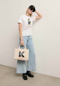 Mulher a usar óculos de sol, boné preto, t-shirt branca do Donald Duck, jeans largos de tom azul claro, mocassins pretos, e segurando uma tote bag bege do Donald Duck.