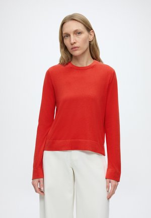 Vrouw met lang blond haar die een rode trui met lange mouwen en witte broek draagt, staand tegen een effen witte achtergrond.