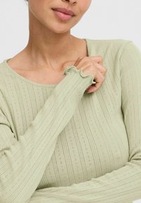 Hele roheline pikakäistega topp ribitud tekstuuriga, millel on peened perforatsioonid. Omab ümmargust kaelust ja volangvarrukaid. Pehme, kehakujuline disain.