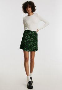 Zwarte rok met groene bloemenpatroon, gecombineerd met een wit overhemd met lange mouwen. Model draagt zwarte schoenen en witte sokken. Eenvoudige, casual look.