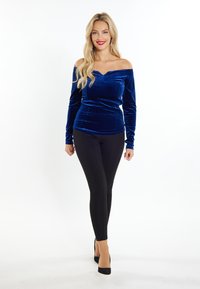 Blauwe fluwelen off-shoulder top met lange mouwen, gecombineerd met zwarte leggings en zwarte hoge hakken, staande tegen een witte achtergrond.