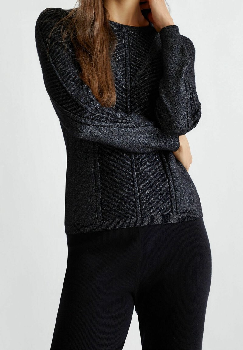Femme portant un pull noir ajusté texturé avec motifs côtes diagonaux et manches longues, associé à un pantalon noir, sur un fond uni.