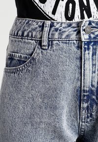 Vaqueros de mezclilla azul claro con una cintura alta, que presentan una textura descolorida, cinco bolsillos y un cierre de botón metálico. Patrón desgastado visible.