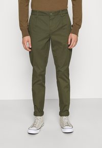Chinos in cotone verde oliva con vestibilità slim, caratterizzati da una chiusura con un singolo bottone e orli arrotolati, abbinati a sneakers alte grigio chiaro.