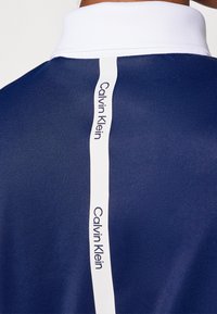 Námořnická polokošile s bílým límečkem, s bílým vertikálním pruhem s nápisem „Calvin Klein“. Hladký materiál, střih na míru.