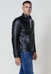 Giacca di pelle nera con un design aderente, caratterizzata da accenti a coste sulle spalle, due tasche con zip e un collo alto. Indossata con jeans blu.