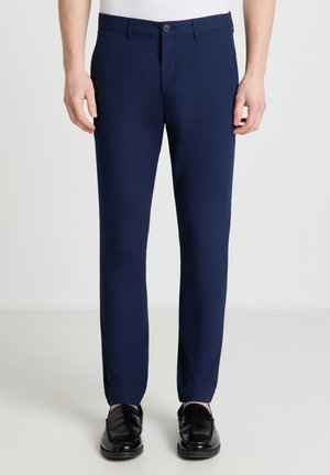 Uomo che indossa pantaloni da abito blu navy slim fit abbinati a mocassini in pelle neri e una maglietta bianca infilata, in piedi su uno sfondo semplice.