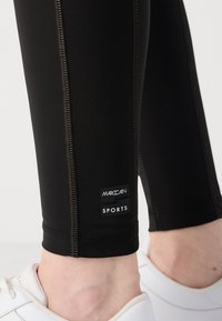 Black sports leggings with contrasting stitching. Features a logo tag on the lower leg. Worn with white athletic shoes.