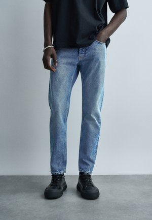 Άνδρας που φορά ανοιχτό μπλε τζιν, μαύρη μπλούζα, μαύρα αθλητικά παπούτσια, με το ένα χέρι στην τσέπη και ένα λευκό βραχιόλι με χάντρες στον καρπό.