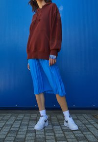 Sudadera oversized color burdeos, falda plisada azul, deportivas blancas con detalles negros. Fondo texturizado en azul, creando un contraste vibrante.