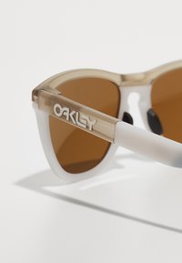 Ochelari de soare unghiulari cu rame transparente de culoare bej, lentile maro și brațe albe cu logo-ul "OAKLEY" scris cu litere îndrăznețe.