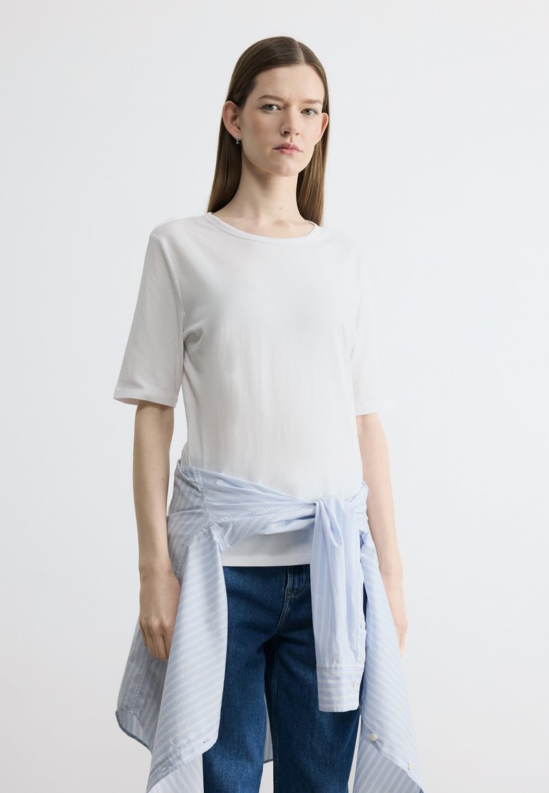 Jeune femme aux longs cheveux bruns portant un T-shirt blanc uni, un jean bleu et une chemise à rayures bleu clair nouée autour de la taille.