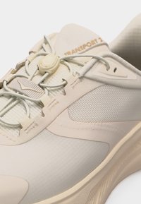 Μπεζ αθλητικό παπούτσι με υφή υφάσματος, ρυθμιζόμενα ελαστικά κορδόνια ασφαλισμένα με πλαστικό κούμπωμα και το λογότυπο "TRANSPORT 2" στη γλώσσα.