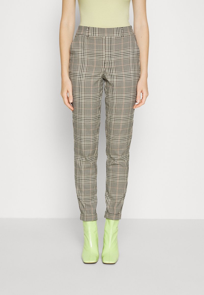 Femme portant un pantalon à carreaux noir et blanc taille haute, un top sans manches jaune pâle et des bottines vert clair, debout sur fond blanc.