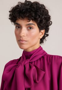 Blusa bordeaux con un dettaglio a fiocco annodato al collo, realizzata in un tessuto morbido e fluido con una sottile texture, scollatura rotonda e maniche lunghe a sbuffo.