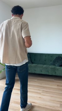 Zelený sametový gauč s prešívaným dizajnom a dvoma textúrovanými vankúšmi. Svetlé drevené podlahy a rastlina v rohu. Osoba v béžovej košeli a džínsach.