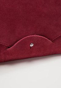 Gros plan sur un rabat de sac en daim rouge avec bord arrondi et petite fermeture bouton argentée sur une surface blanche.