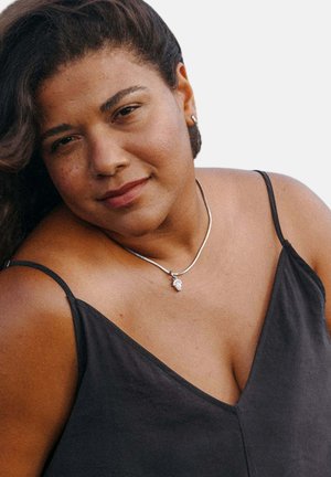 Frau mit mittlerem Hautton und dunklen, lockigen Haaren, die ein schwarzes ärmelloses Oberteil, eine silberne Halskette und kleine Ohrringe trägt und sanft in die Kamera blickt.