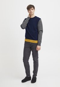 Férfi színblokkkal ellátott pulóver: tengerészkék test, szürke ujjak és sárga bordázott alja. Szürke, slim fit farmerrel és fekete cipővel párosítva.