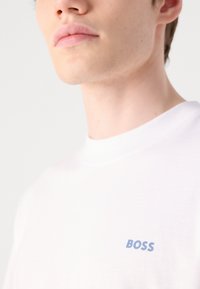 Bílý bavlněný tričko s malým modrým logem "BOSS" na levé straně. Měkká textura a kulatý výstřih, detailní pohled na horní část oděvu.