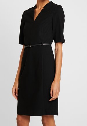 Schwarzes knielanges Kleid mit kurzen Ärmeln, V-Ausschnitt und schlankem schwarzem Gürtel in der Taille, getragen von einer stehenden Person mit nackten Armen.