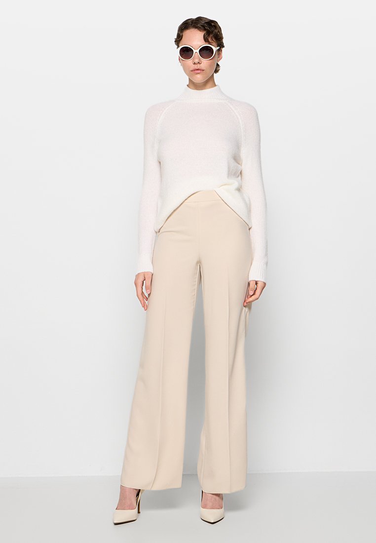 Suéter blanco de gran tamaño con cuello alto, combinado con pantalones de pierna ancha beige. Los accesorios incluyen gafas de sol redondas blancas y tacones beige de punta.