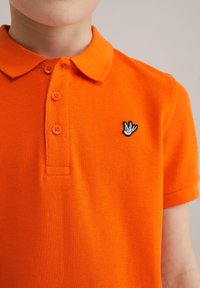 Pomarańczowa koszulka polo z teksturowanej tkaniny, z kołnierzykiem, trzema guzikami oraz czarno-białym haftowanym logo na piersi.