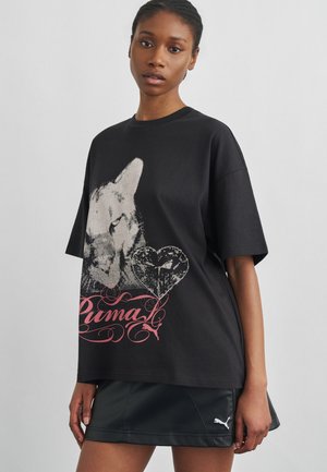 Mlada žena nosi crnu oversized majicu s velikom grafikom mačke i ružičastim tekstom "Puma", u kombinaciji s crnom suknjom s logom Puma.