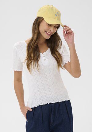 Femeie cu păr lung, purtând o șapcă galbenă cu inimioare albastre, un top tricotat alb cu mâneci scurte și pantaloni albastru închis, zâmbind cu ochii închiși.