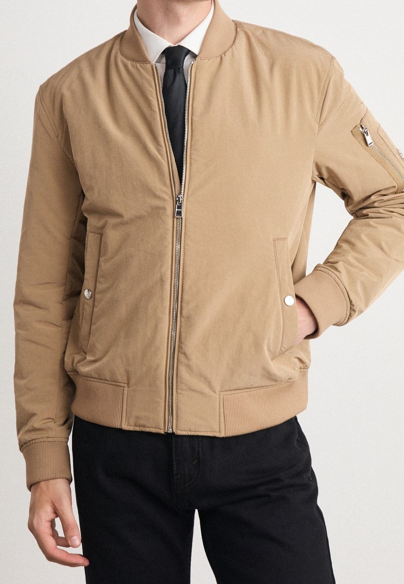Tan bomberjacka med dragkedja, ribbade ärmslut och nederkant, sidofickor och en dekorativ dragkedja på ärmen. Slät, matt textur.