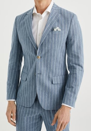 Homme portant un costume bleu à fines rayures, une veste à un bouton, une chemise blanche et un mouchoir de poche à motifs.