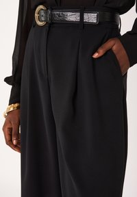 Zwarte broek met hoge taille en plooien, gecombineerd met een glanzende zwarte riem met een gouden ronde gesp, afgebeeld met een hand in de zak.