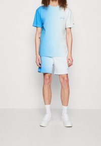 Světle modré tričko s krátkým rukávem a vyšitým logem, v kombinaci s odpovídajícími šortkami. Texturovaná tkanina, casual fit, bílé tenisky.