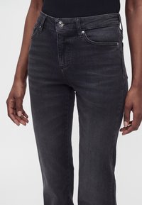 Gros plan d'un jean droit gris foncé porté par une personne à la peau foncée, montrant les poches avant et la fermeture par boutons.