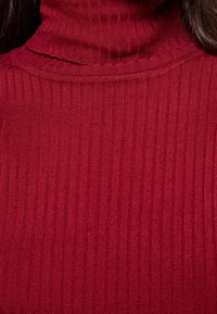 Pull à col roulé rouge à côtes, fabriqué en tissu doux, avec un col montant et une surface texturée de lignes verticales.