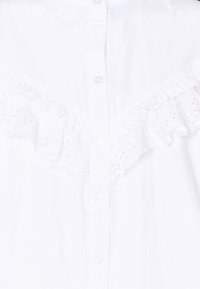 Chemise blanche à boutons sur le devant, avec des accents à volants et des détails en dentelle brodée le long de l'encolure. Tissu en coton lisse.