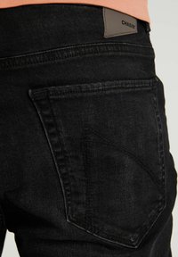Zwarte denim jeans met een gestructureerde stof, schuin achterzak en een leren label met het "CHASIN'" logo op de tailleband.