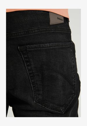Jeans de denim negro con tela texturizada, bolsillo trasero en ángulo y una etiqueta de cuero con el logo "CHASIN'" en la cinturilla.